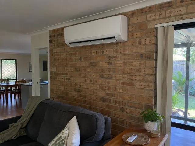 Split system indoor unit on a brick wall above a lounge room sofa in a home in Oxenford