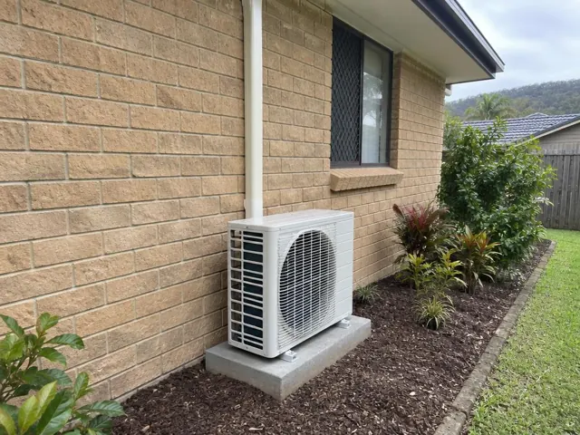 Outdoor air conditioner condenser on a concrete pad beside a brick house and garden in Maudsland